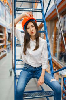 Female customer in helmet sitting on the stairs in hardware store. Buyer look at the goods in diy shop, shopping in building supermarket