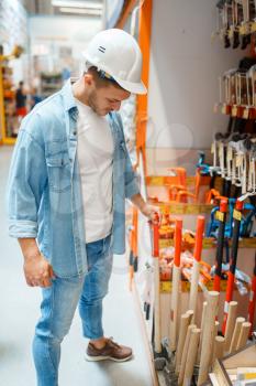 Male buyer in white helmet choosing hammer in hardware store. Customer look at the goods in diy shop, shopping in building supermarket