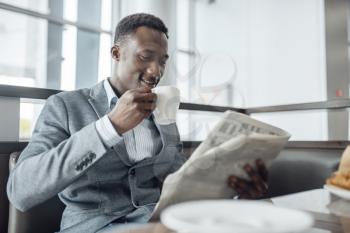 Young black businessman with newspaper having lunch in office cafe. Successful business person drinks coffee in food-court, black man in formal wear