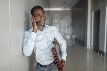 Black businessman with briefcase talking by phone in office hallway. Successful business person negotiates in corridor, black man in formal wear