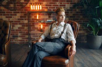 Elegance business woman in strict clothes poses on leather couch in studio, retro fashion, gangster style, mafia.