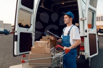 Deliveryman in uniform holds cart with boxes at the car with parcels, delivery service. Man standing at cardboard packages in vehicle, male deliver, courier or shipping job