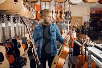 Bearded young guy choosing acoustic guitar in music store. Assortment in musical instruments shop, male musician buying equipment