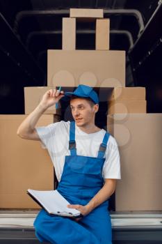 Deliveryman in uniform, parcels in the car on background, cargo, delivery service. Man standing at cardboard packages in vehicle, male deliver, courier or shipping job