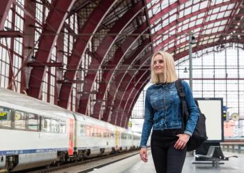 Female tourist with backpack waiting for train on railway station platform, travel in Europe. Transportation by european railroads, comfortable tourism and travelling