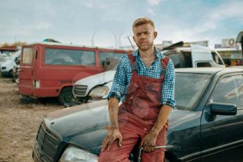 Male repairman poses on car junkyard. Auto scrap, vehicle junk, automobile garbage, abandoned, damaged and crushed transport