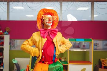 Funny clown in star glasses poses in children's area. Birthday party celebrating in playroom, baby holiday in playground. Childhood happiness, childish leisure, entertainment with animator