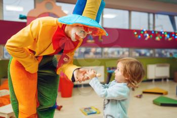 Funny clown gives out lollipop to happy little girl, friendship forever. Birthday party celebrating in playroom, baby holiday in playground. Childhood happiness, childish leisure