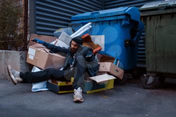 Dirty drunk beggar lies in garbage at the trashcan on city street. Poverty is a social problem, homelessness and loneliness, alcoholism and drunk addiction, urban lonely