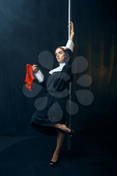 Sexy perverse nun in a cassock dances on a pole with red panties in hands, vicious desires. Corrupt sister in the monastery, sinful religious people, attractive sinner