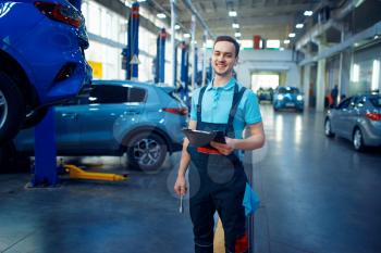 Repairman with a checklist stands at vehicle on lift, car service station. Automobile checking and inspection, professional diagnostics and repair