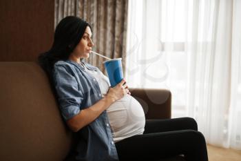 Pregnant woman with belly sitting on sofa and drinks juice from a large glass at home. Pregnancy, gluttony in prenatal period. Expectant mom resting in living room