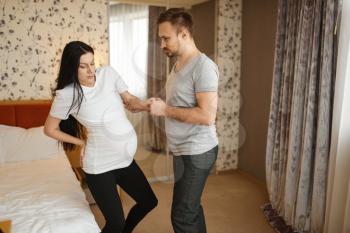 Husband helps his pregnant wife with belly at home, bedroom interior on background. Pregnancy, prenatal period. Expectant mom and dad are resting on sofa, health care
