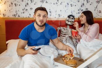 Husband watching TV, wife feeding human skeleton in the bed, bad relationship. Couple having a problems, family quarrel, conflict of married man and woman