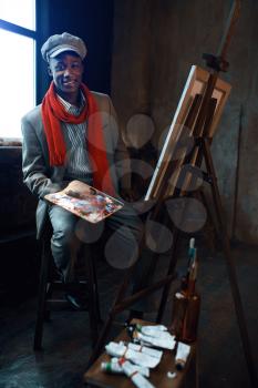 Fashionable male painter with palette poses at easel in art studio. Artist draws at his workplace, creative master works in workshop