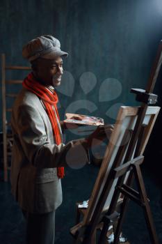 Fashionable male painter with palette poses at easel in art studio. Artist draws at his workplace
