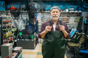 Fisherman tries on rubber jumpsuit in fishing shop, hooks and baubles on background. Equipment and tools for fish catching and hunting, accessory choice on showcase in store
