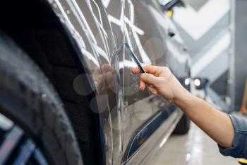 Specialist cuts protection film on car surface. Installation of coating that protects the paint of automobile from scratches. New vehicle in garage, tuning procedure