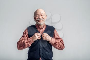 Smiling elderly man with mustache puts on a jacket, grey background. Cheerful mature senior looking at camera in studio