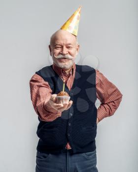 Smiling elderly man in party cap shows cake with candle, grey background. Cheerful mature senior looking at camera in studio