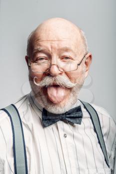 Portrait of elderly man in a bow tie and glasses showing his tongue, grey background. Mature senior looking at camera in studio