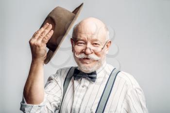Elderly man in a bow tie and glasses takes off his hat, grey background. Mature senior looking at camera in studio