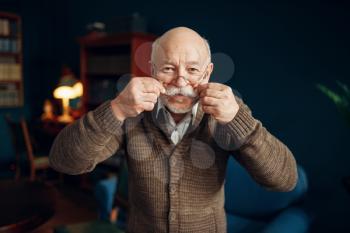 Cheerful elderly man shows his moustache in home office. Bearded mature senior poses in living room, old age businessman