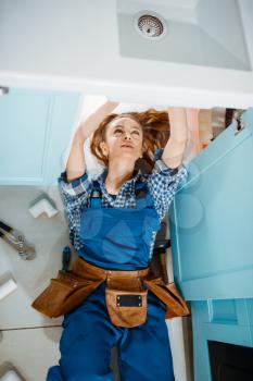 Female plumber in uniform lying on the floor in the kitchen, top view. Handywoman with toolbag repair sink, sanitary equipment service at home