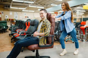 Happy young couple rides on office chair in furniture store showroom. Man and woman looking samples for bedroom in shop, husband and wife buys goods for modern home interior