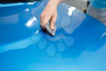 Car wrapping, mechanic with squeegee installs protective vinyl foil or film on vehicle hood. Worker makes auto detailing. Automobile paint protection coating, professional tuning