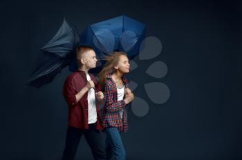 Little boy and girl with umbrellas in studio, wind effect. Children with developing hairs, kids isolated on dark background, child emotion