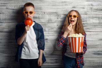Little boy and girl in sunglasses holds popcorn and big lollipop in studio. Children and sweets, kids isolated on wooden background, child photo session