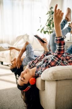 Two happy women enjoys listening to music upside down at home. Pretty girlfriends in earphones relax in the room, sound lovers resting on couch, female friends leisures together