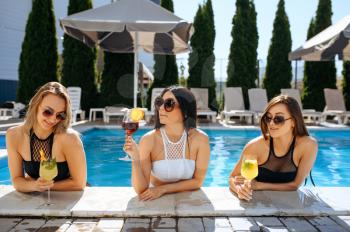 Sexy women drinks coctails in the pool outdoors. Beautiful girls in sunglasses relax at the poolside in sunny day, summer holidays of attractive girlfriends