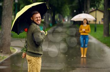 Love couple with umbrellas meets in park, rainy day. Man with red rose waiting for his woman on walking path, wet weather in alley
