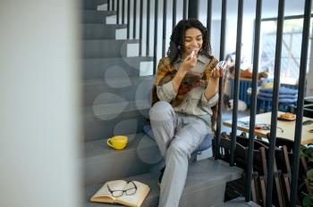 Smiling female student with lipstick sitting on the steps in library cafe. Woman with cup of coffee, opened book and glasses on background, education and knowledge. Girl studying in campus