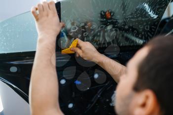 Male worker installs wetted car tinting, tuning service. Mechanic applying vinyl tint on vehicle window in garage, tinted glass