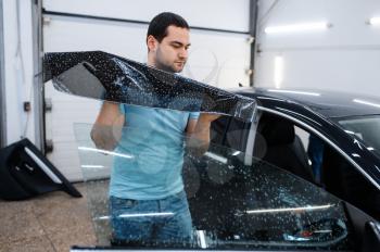 Male specialist installs wetted car tinting, tuning service. Mechanic applying vinyl tint on vehicle window in garage, tinted automobile glass