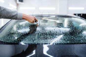 Male worker with squeegee cleans car for tinting, tuning service. Mechanic applying vinyl tint on vehicle window in garage, tinted automobile glass