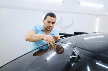 Male wrapper cuts car tinting, tuning service. Worker applying vinyl tint on vehicle window in the garage