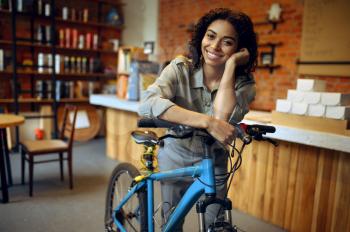 Smiling female student poses with bicycle in cafe. Woman with cycle in coffeehouse. Girl leisures in campus cafeteria