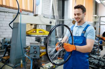 Worker in uniform holds bicycle wheel near machine tool on factory. Bike rims and spokes assembly in workshop, cycle parts installation, , modern technology