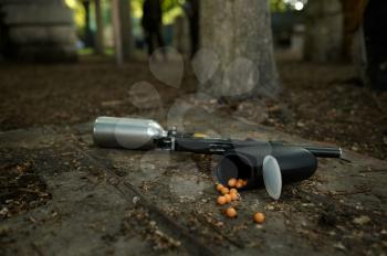 Paintball gun and marker balls on the ground closeup, nobody, playground in the forest on background. Extreme sport outdoors, pneumatic weapon and paint bullets, military team game concept