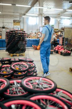 Bicycle factory, worker at the stack of bike wheels. Male mechanic in uniform installs cycle parts, assembly line in workshop, industrial manufacturing