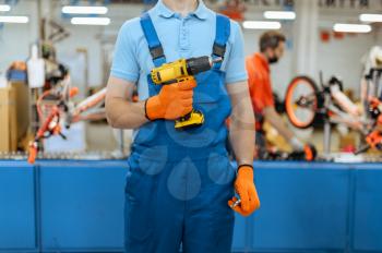 Bicycle factory, worker holds electric screwdriver at bike assembly line. Male mechanic in uniform installs cycle parts in workshop, industrial manufacturing
