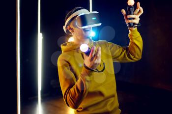 Male gamer plays the game using virtual reality headset and gamepad in luminous cube. Dark playing club interior, spotlight on background, VR technology, 3D vision