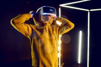 Young gamer plays the game using virtual reality helmet and gamepad in luminous cube. Dark playing club interior, spotlight on background, VR technology with 3D vision