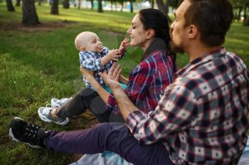 Mother, father and little baby rests in summer park. Mom and dad with male kid on lawn, picnic with child on plaid in the forest, family happiness