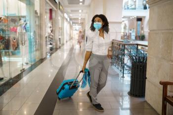Young woman with suitcase walking in mall. Female traveler with luggage in shopping center, passenger with bag in tax free market. Girl in mask with baggage