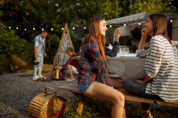 Young friends relax on picnic at camping in the forest. Youth having summer adventure on rv, camping-car on background. Two couples leisures, travelling with trailer